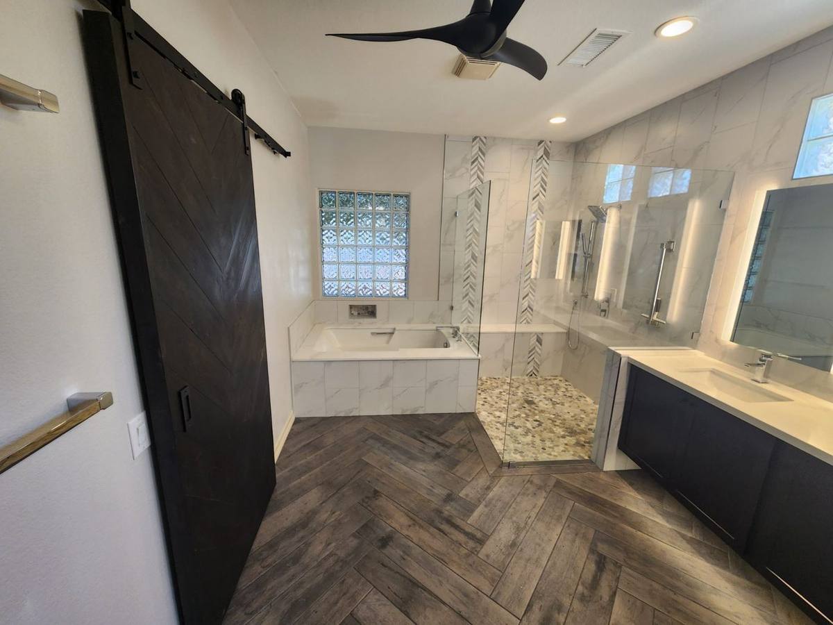 Modern primary bathroom featuring herringbone wood-look tile flooring, dark barn door, soaking tub, and marble walk-in shower